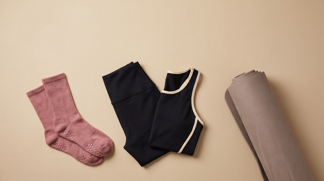 Flat-Lay aus Dusty-Rose-Grip-Socken, dunklem Pilates-Outfit mit Cream-Kontrast und gerollter Matte auf Sand-Beige-Hintergrund